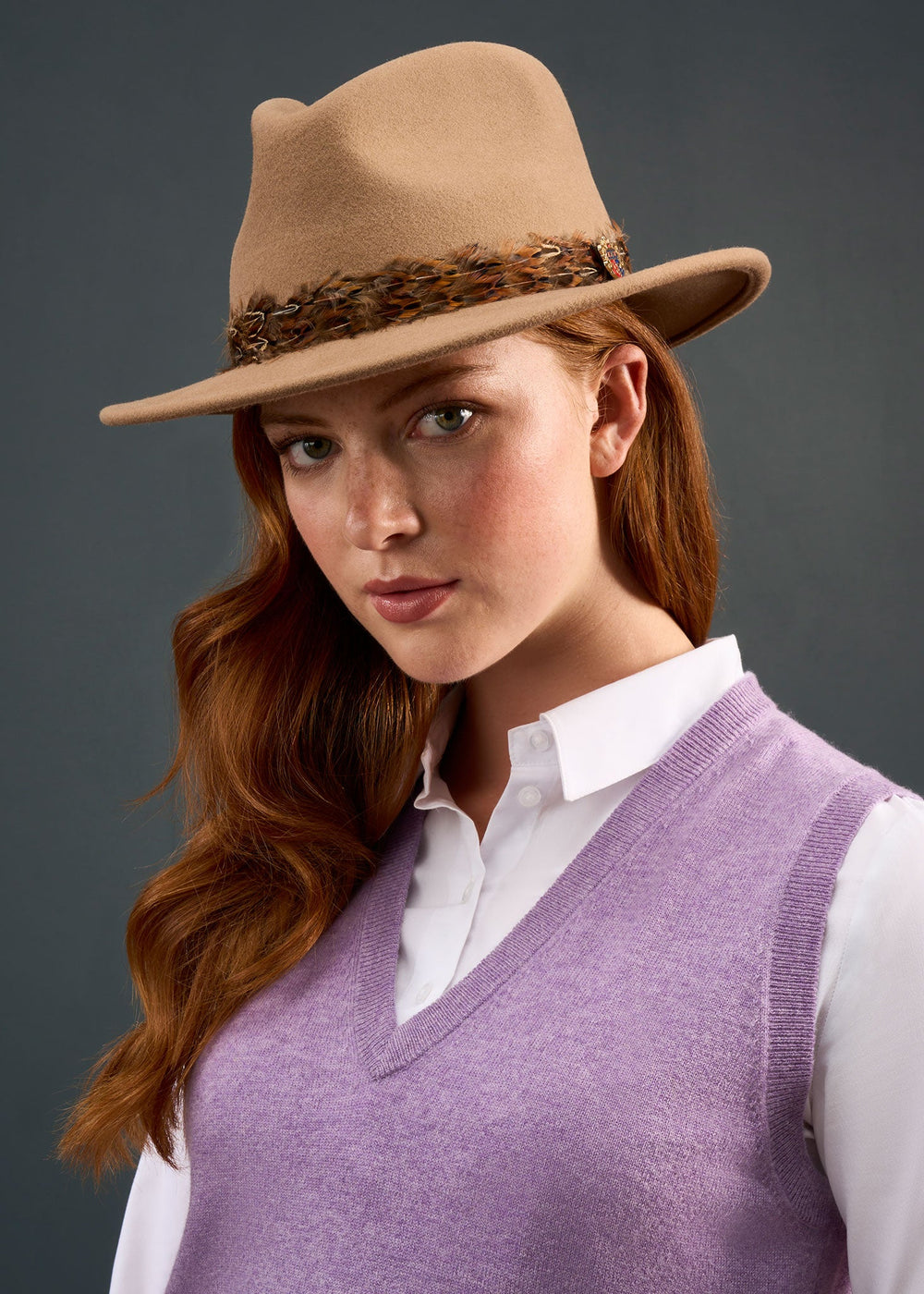 Woman with long red hair in an Erin Wool & Cashmere V-Neck Slipover, brown hat, and lavender vest against a gray backdrop.