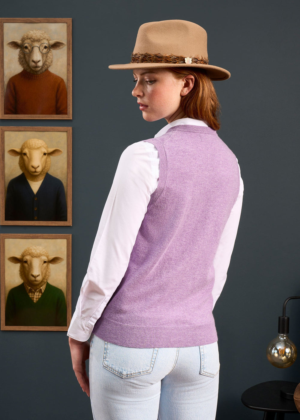 Woman in hat and purple vest stands by 3 sheep portraits, wearing the Erin Wool & Cashmere V-Neck Slipover.