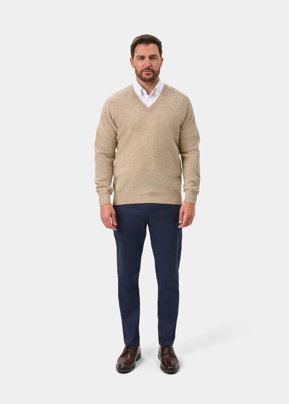 Man wears Hampshire Lambswool V-Neck Jumper in Linen over a white shirt, navy pants, and brown shoes.