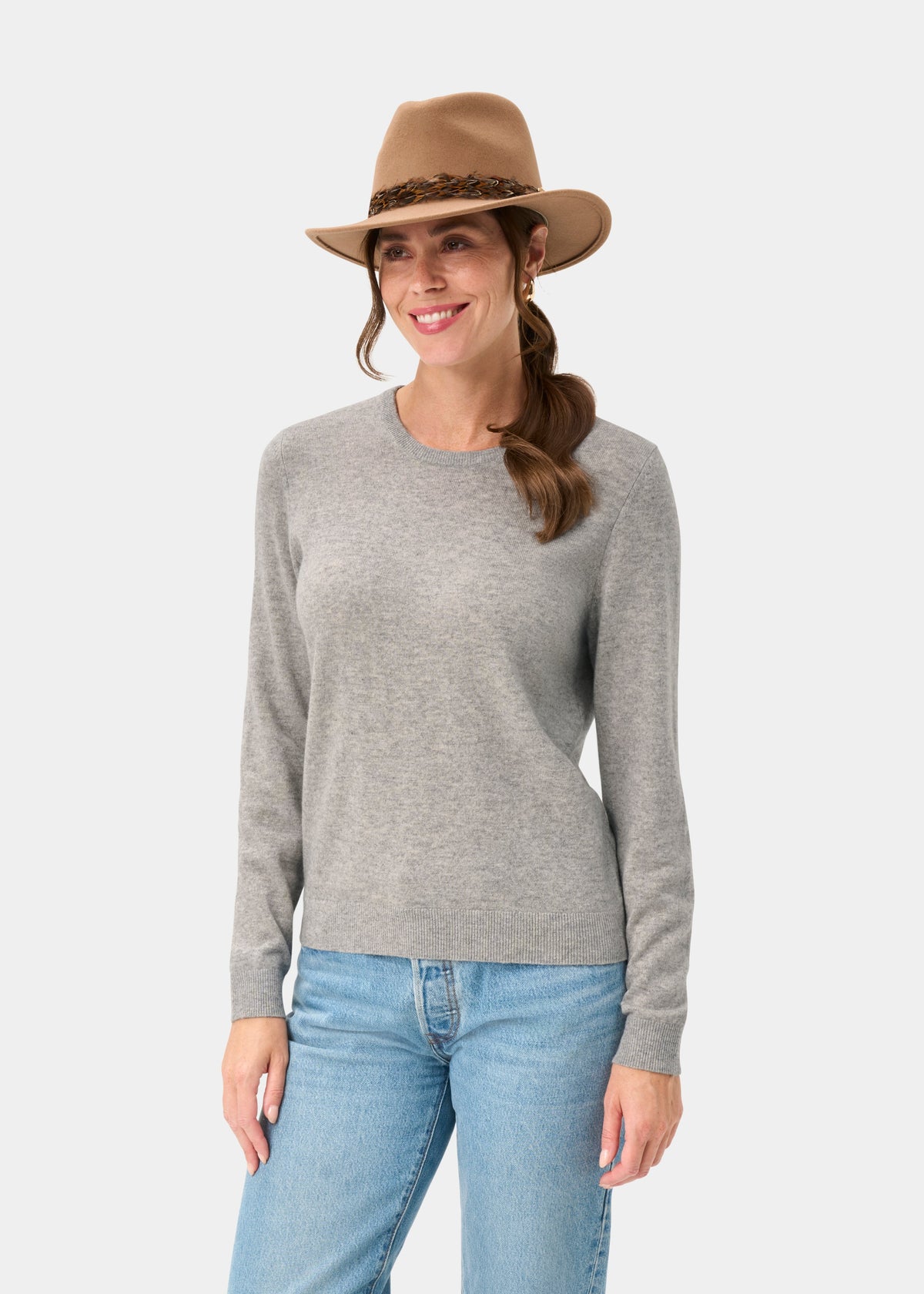 Woman in Ivy Wool & Cashmere Crew Neck Jumper in Husky, blue jeans, and brown hat smiling on a white background.