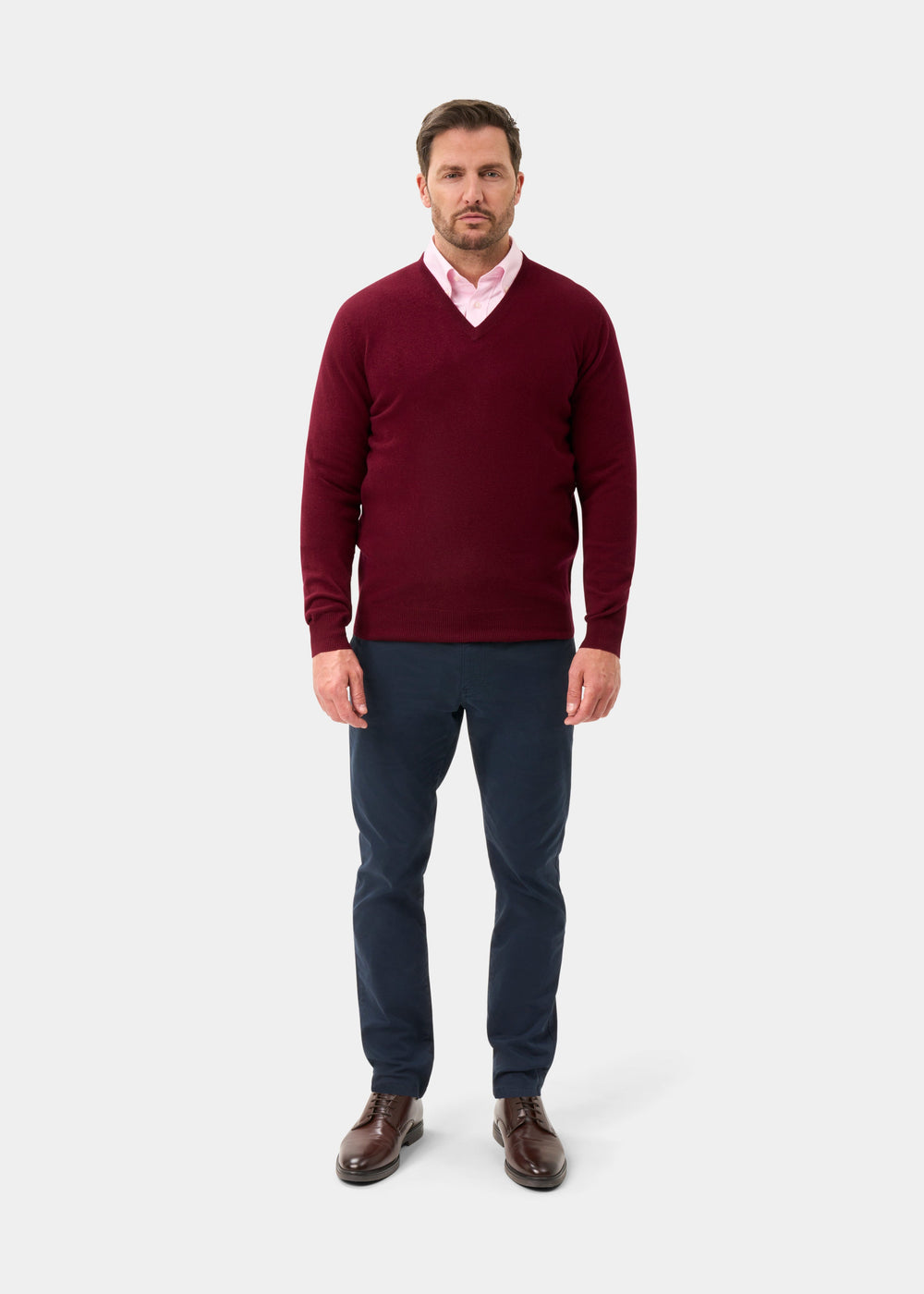 Man wears Kilsyth Lambswool V-Neck Jumper in Garnet, pink shirt, navy pants, and brown shoes on white background.