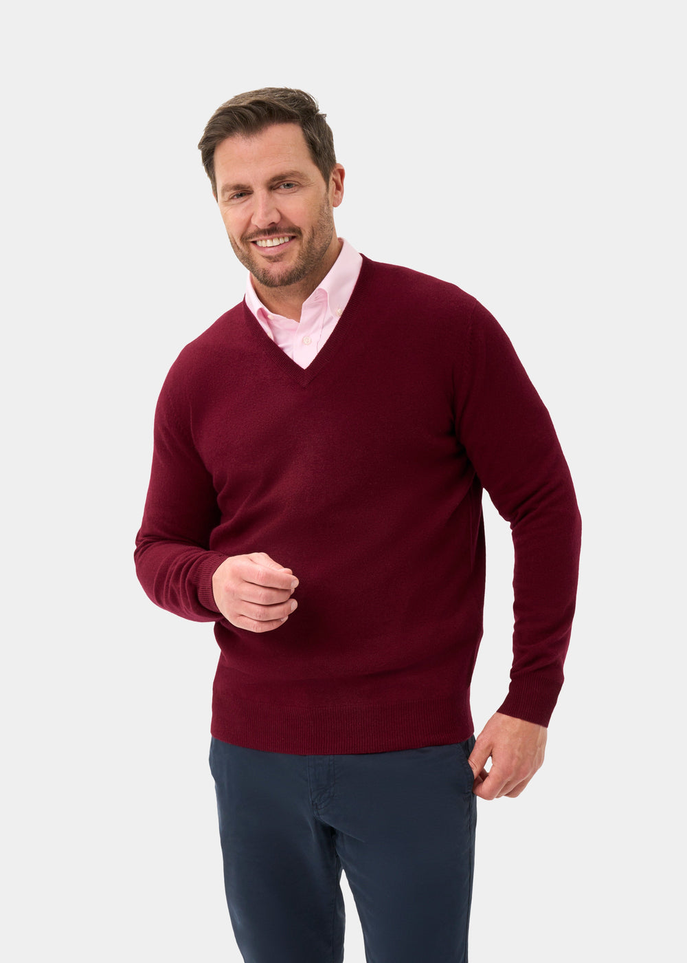 Man in Kilsyth Lambswool V-Neck Jumper in Garnet over pink shirt and navy pants, smiling against light background.