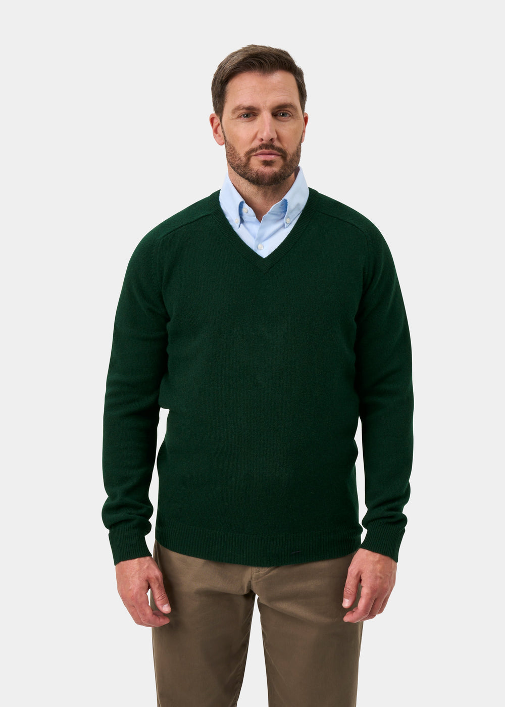 Man wears Kilsyth Lambswool V-Neck Jumper in Mallard over a blue shirt and khaki pants, standing against white background.