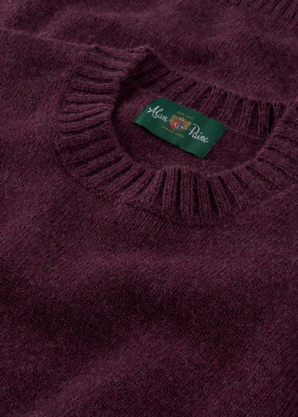 A close-up of the Kinnadie Shetland Wool Crew Neck Jumper in Elderberry shows its ribbed collar and green label.