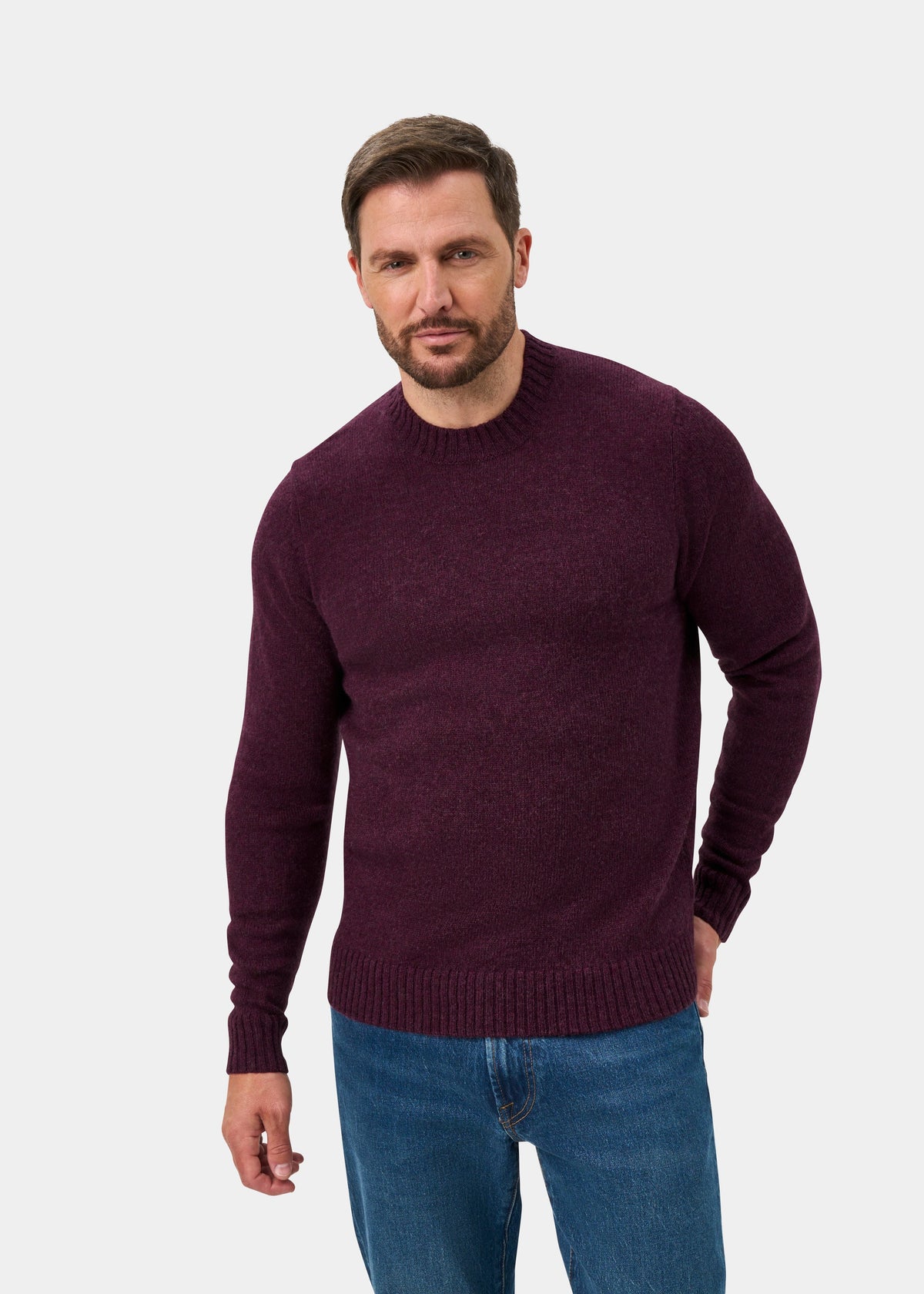 Man in a Kinnadie Supersoft Shetland Wool Crew Neck Jumper in Elderberry and blue jeans stands against a gray background.