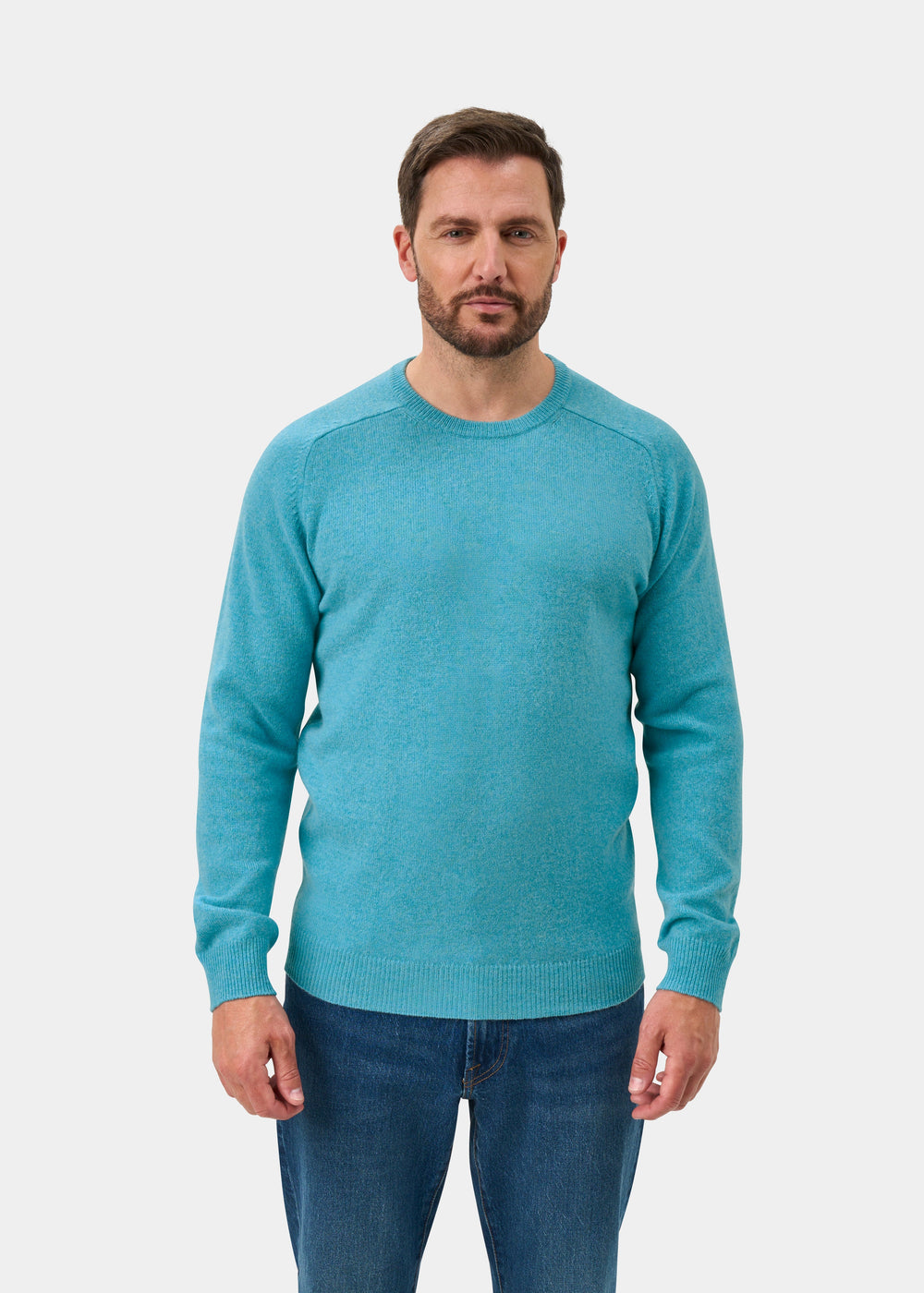 Man wears Lenzie Lambswool Crew Neck Jumper in Topaz, regular fit, with blue jeans, facing forward on white background.