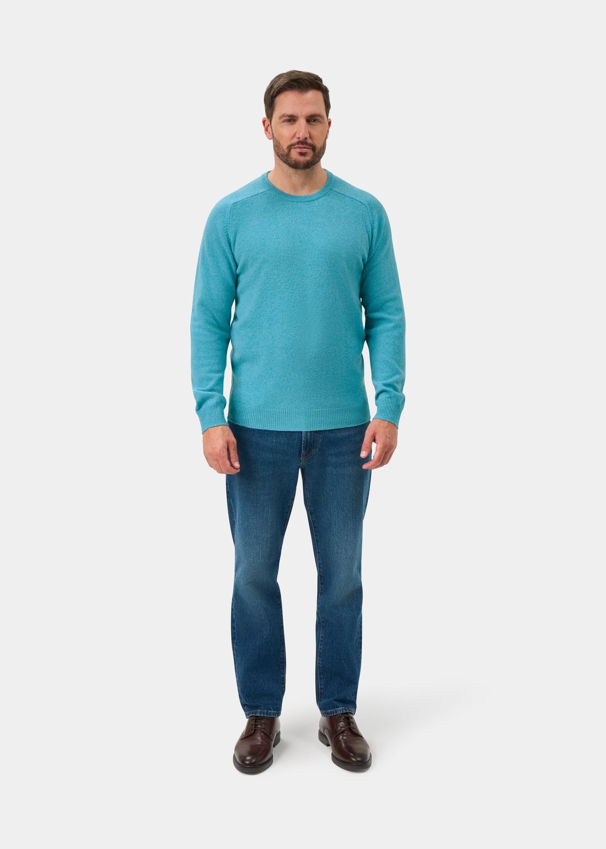 Man in Lenzie Lambswool Crew Neck Jumper in Topaz, blue jeans, and brown shoes on a white background.