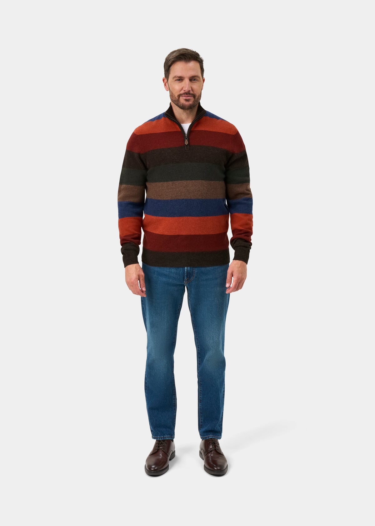A man wearing a multi-colored horizontally striped quarter-zip sweater, blue jeans, and brown leather shoes, standing against a light gray studio background.