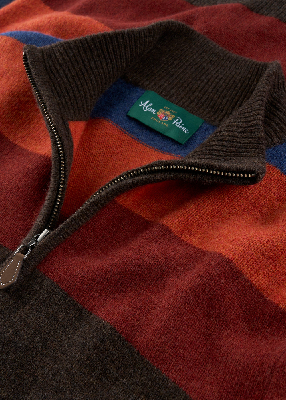 Close-up of a knit quarter-zip sweater with a dark brown ribbed collar and zipper, showing a green Man & Planter brand label. The sweater features a warm-toned plaid pattern with rust red, orange-brown, dark brown, and blue blocks.