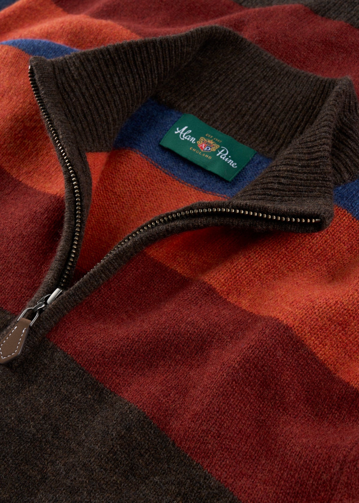 Close-up of a knit quarter-zip sweater with a dark brown ribbed collar and zipper, showing a green Man & Planter brand label. The sweater features a warm-toned plaid pattern with rust red, orange-brown, dark brown, and blue blocks.