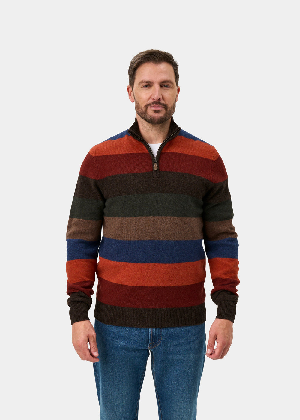 A man wearing a horizontally striped quarter-zip sweater in shades of rust red, dark brown, olive green, and blue, paired with blue jeans, against a plain light gray background.