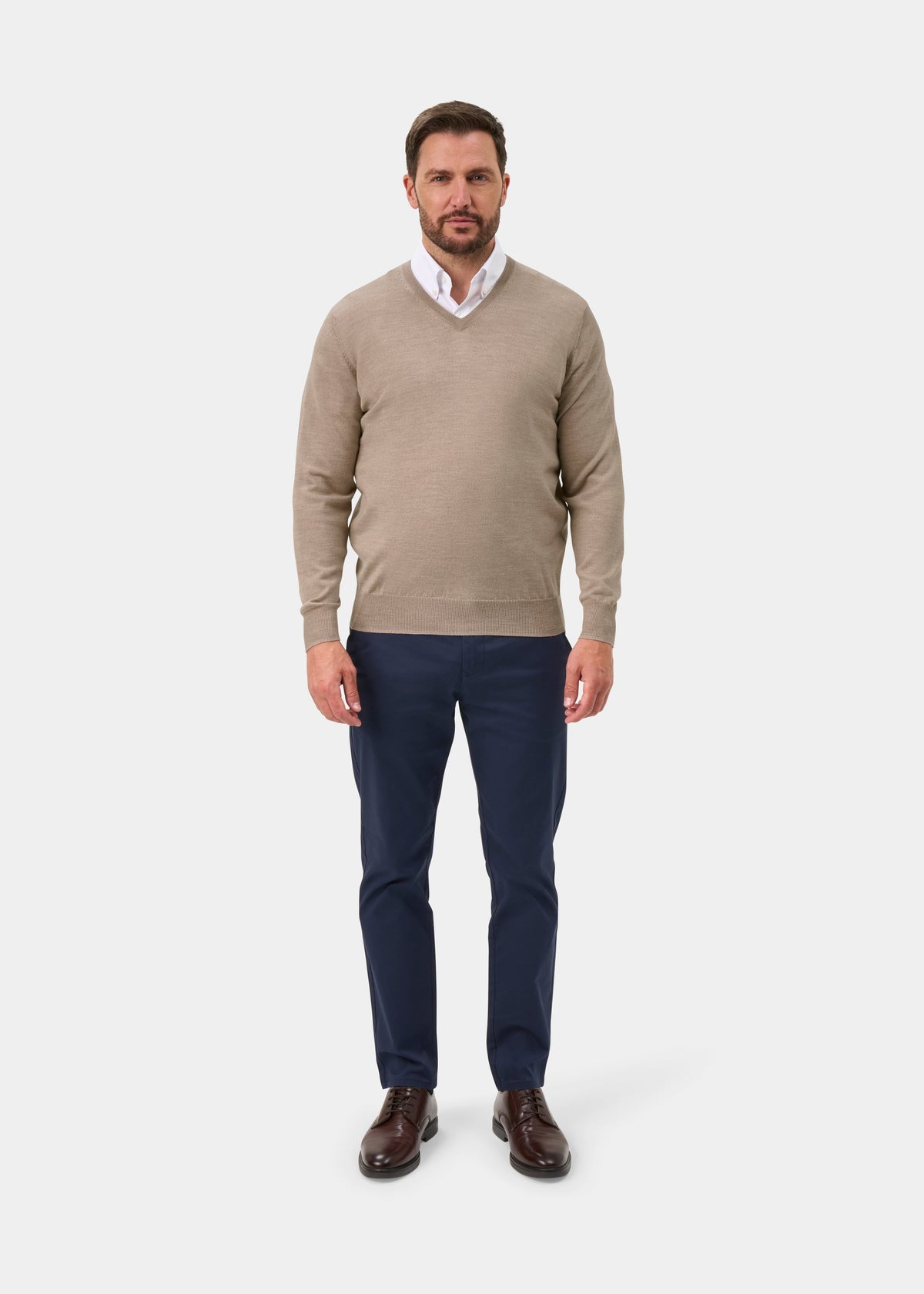A man wearing a beige v-neck sweater over a white collared shirt, navy blue trousers, and brown leather shoes, standing against a light gray studio background.