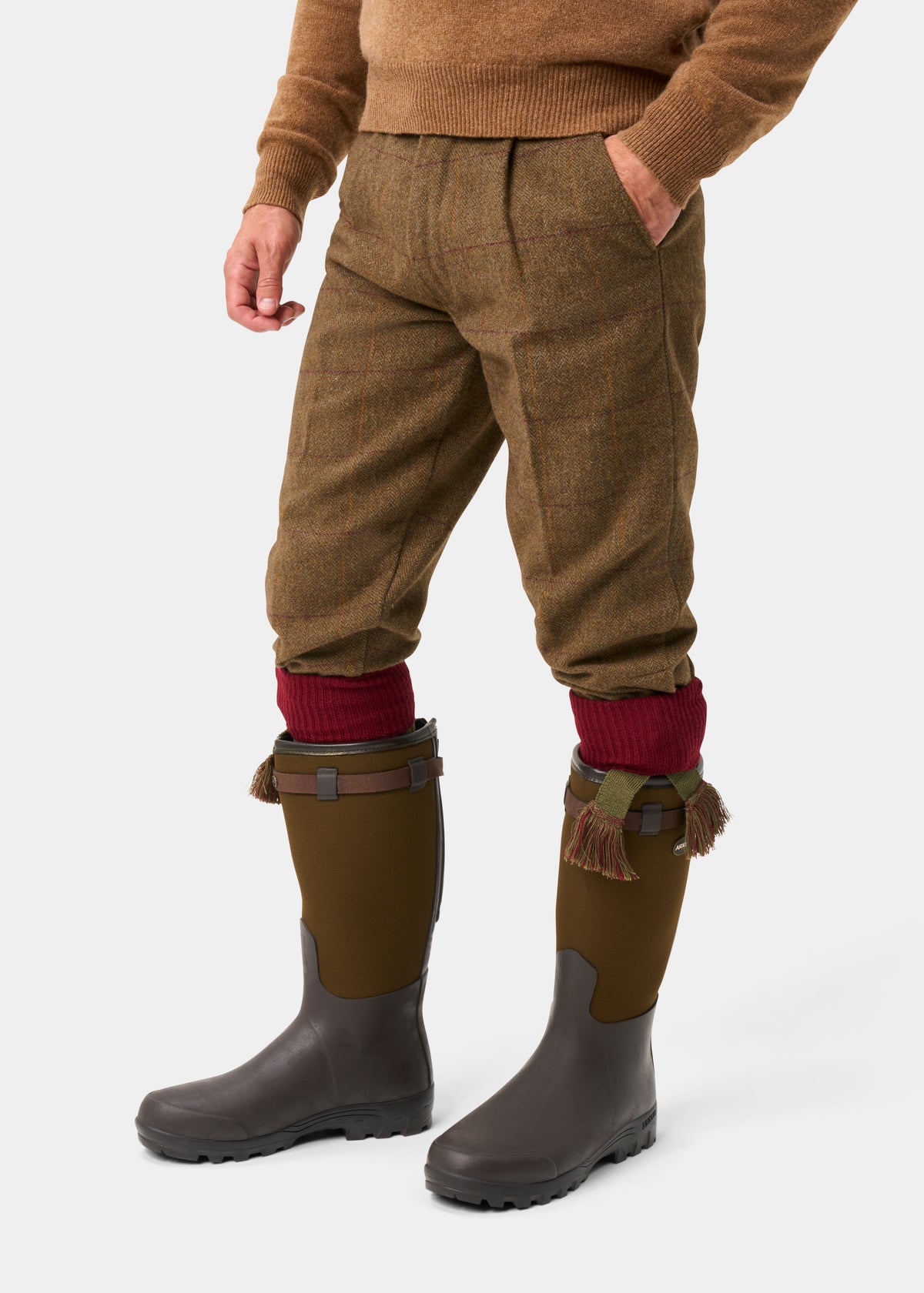 A man wears Rutland Men’s Tweed Water Repellent Shooting Breeks in Birch, red knit socks, and olive green rubber boots.